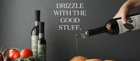 A hand holds a bottle of olive oil with a metal pour spout, drizzling oil into a pan. Two dark olive oil bottles and fresh vegetables like tomatoes and garlic are arranged on a dark gray background, with the text 'DRIZZLE WITH THE GOOD STUFF.